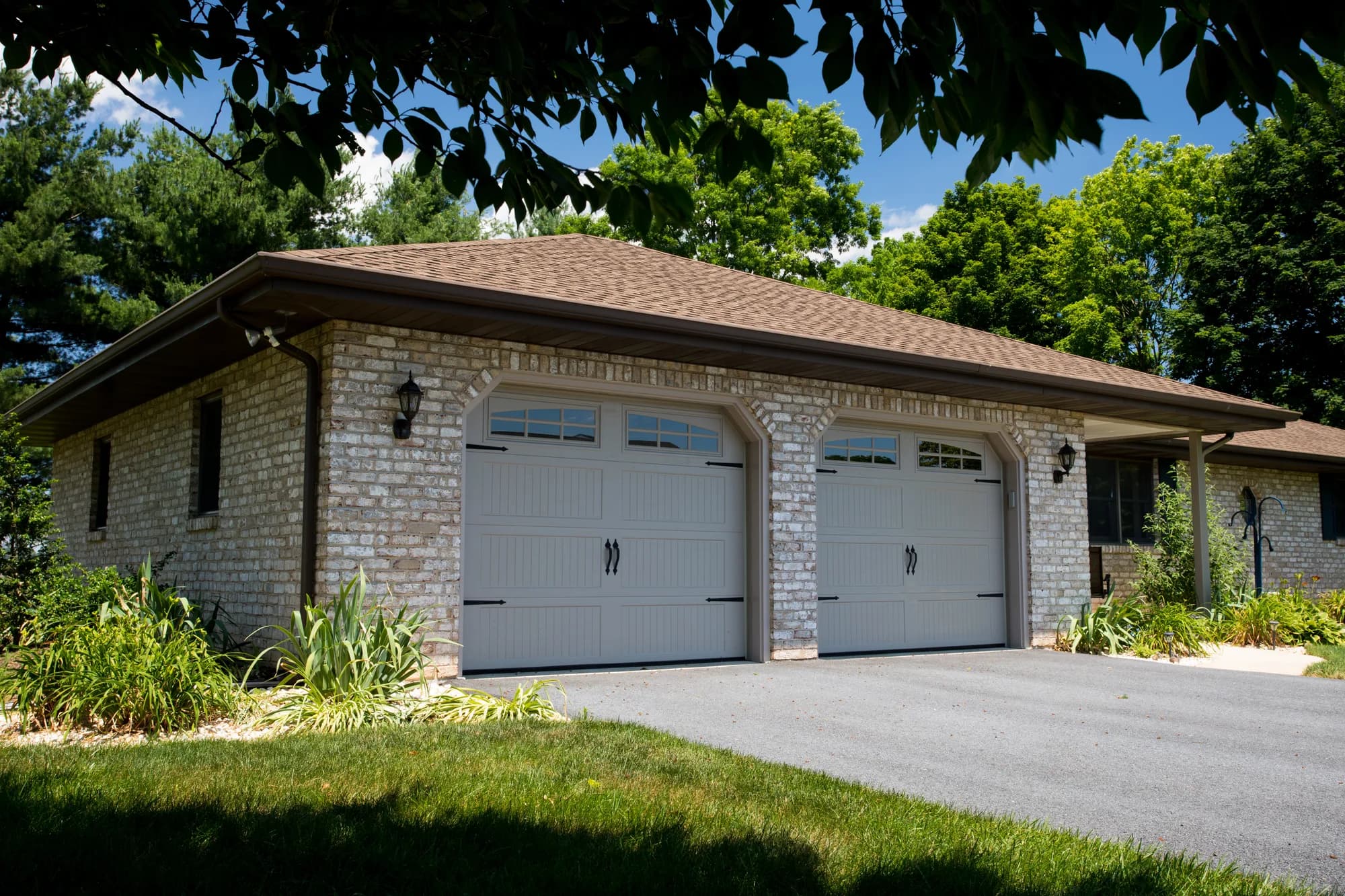 Two taupe carriage-style Haas garage doors with arched top windows and black hardware on a brick garage