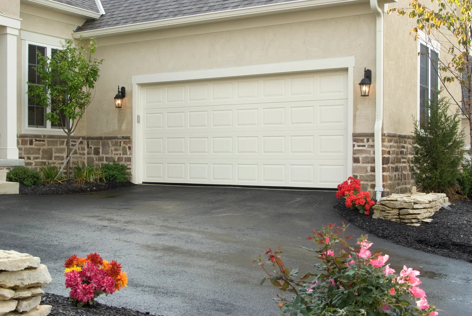 Almond short raised-panel Haas garage door on a stucco home with a stone base and coach lanterns
