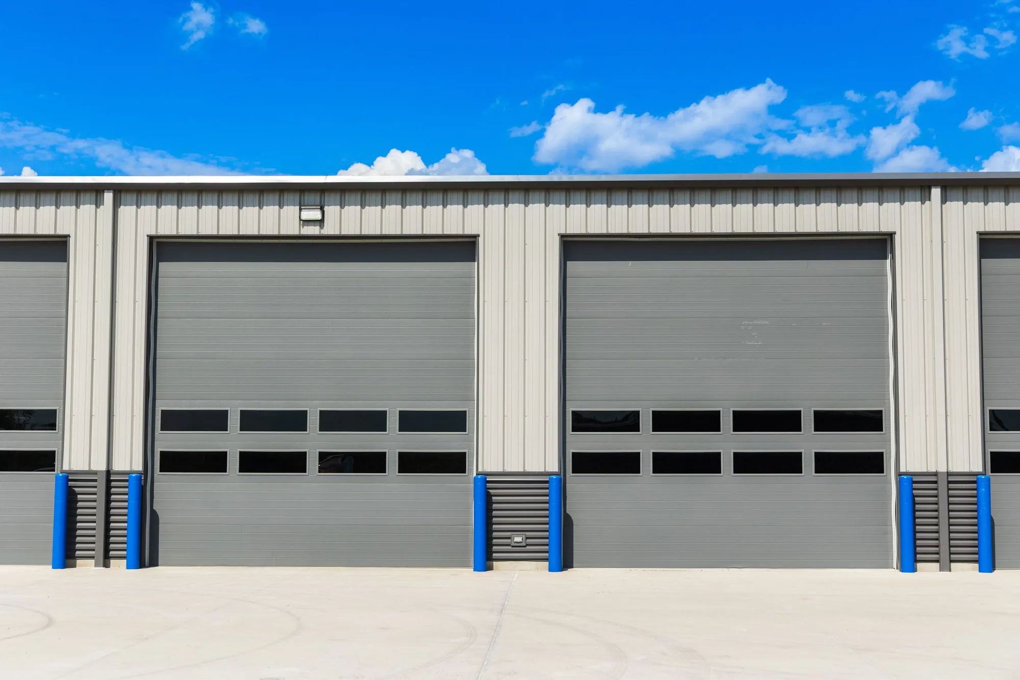 Row of grey sectional steel doors with vision panels and blue bollards on a tan steel commercial building