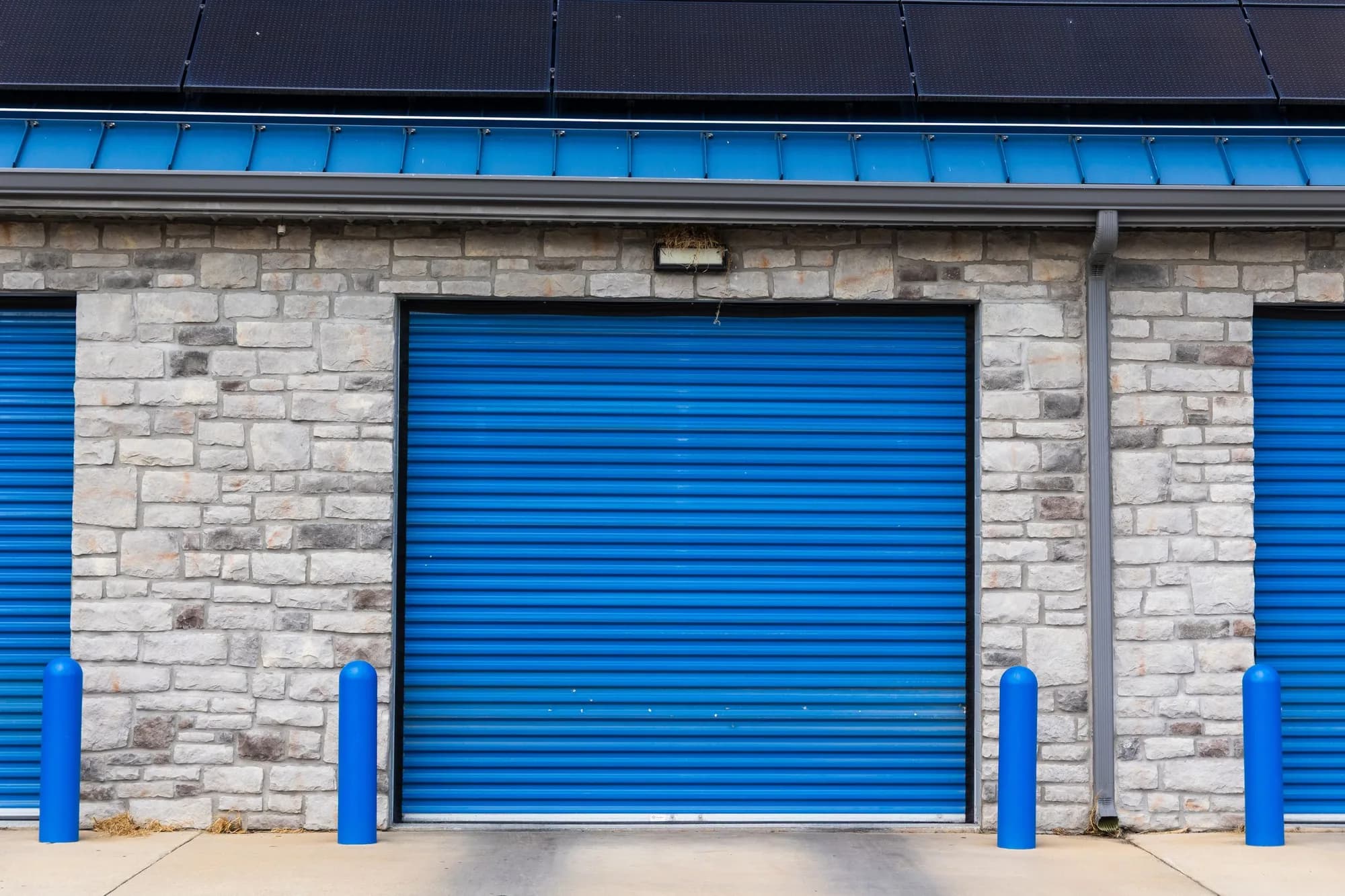 Blue rolling steel commercial door with blue bollards on a stone-faced building
