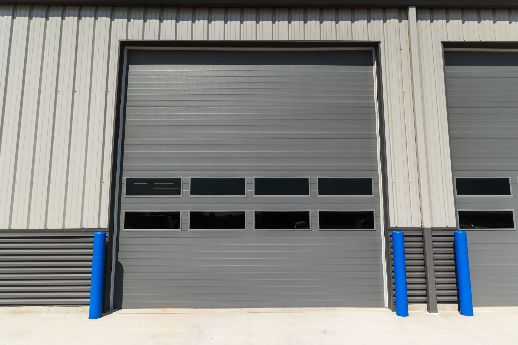Grey sectional steel commercial door with a row of vision panels between blue bollards