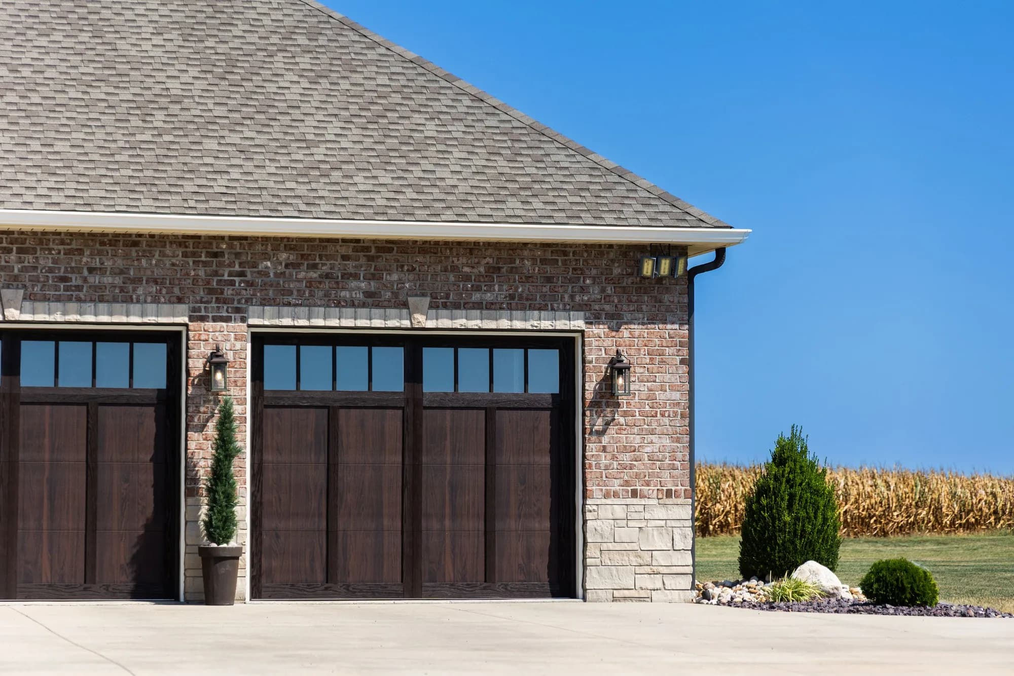 Brick home with two dark woodtone carriage garage doors and a stone base, a cornfield behind, seen from the driveway