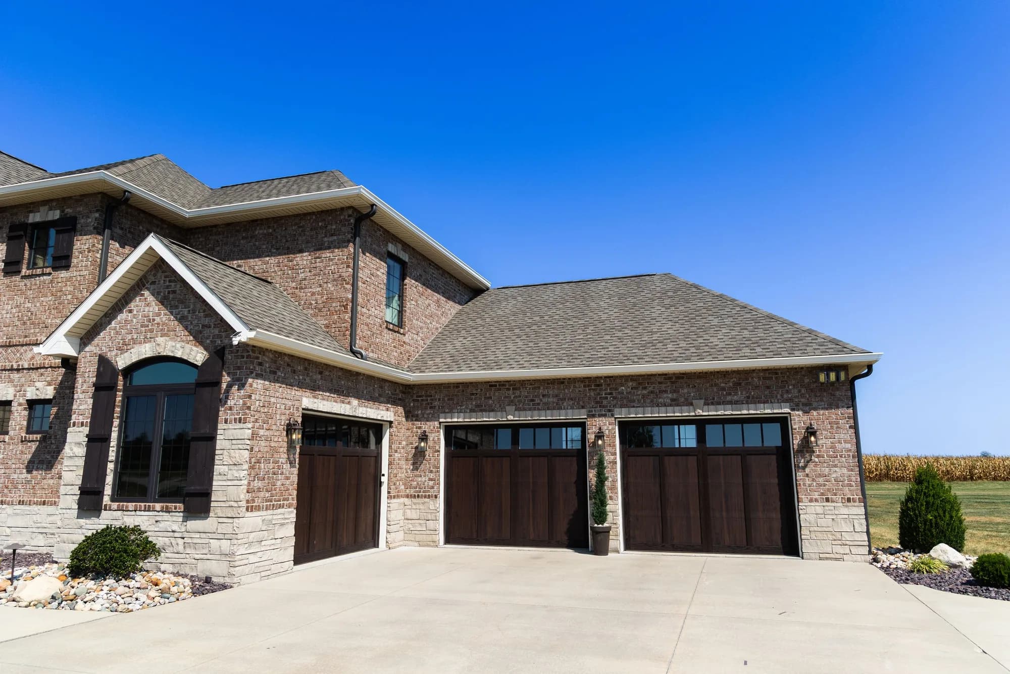 Brick home with two dark woodtone carriage garage doors and a cornfield behind, seen from the driveway