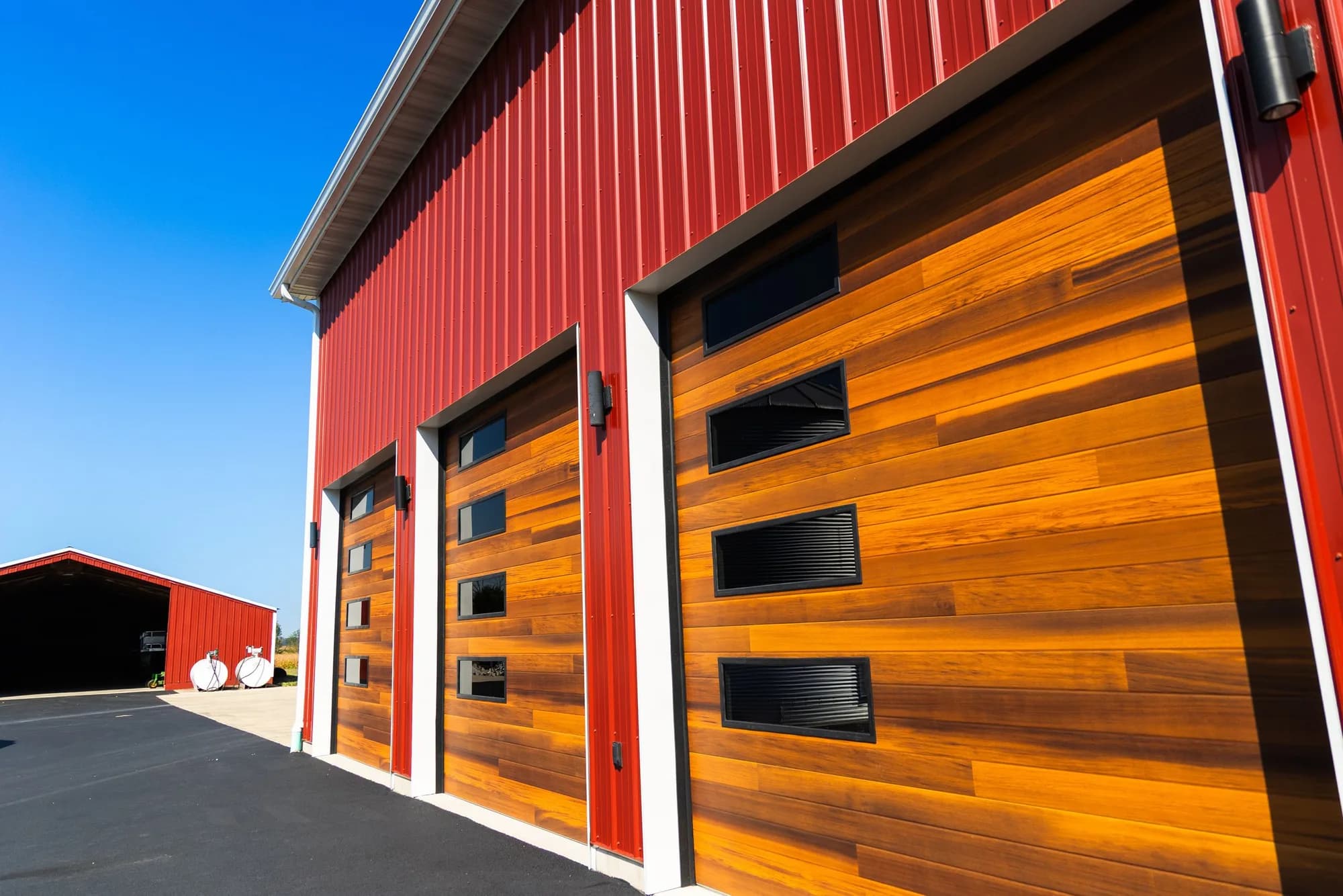 Cedar-tone plank garage doors with stacked windows on a red steel shop, seen from the drive