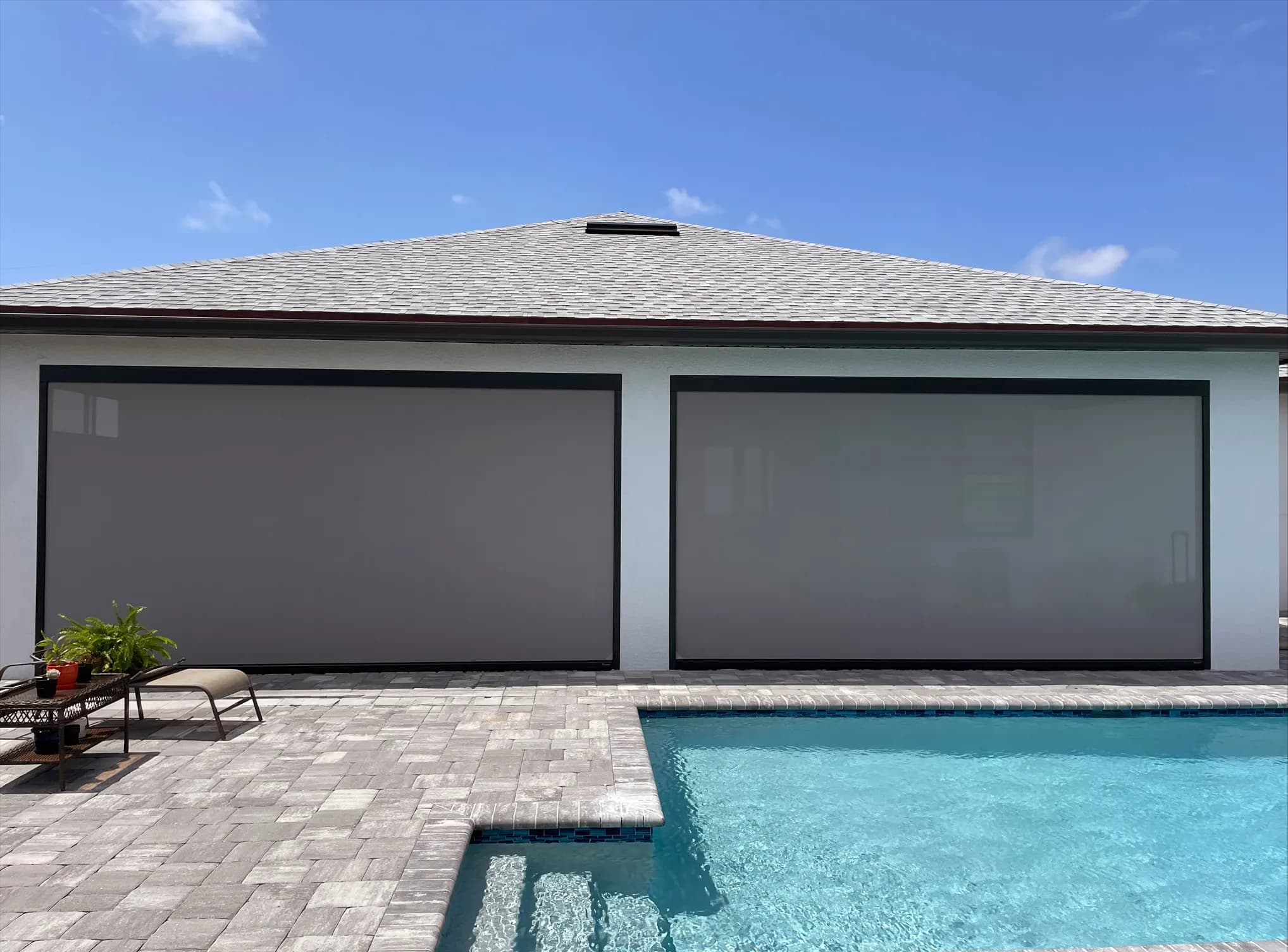 Two wide grey retractable screens lowered across the back of a white stucco house, with a paver patio, a lounge chair, and a pool in front